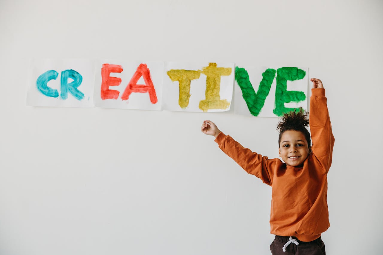 about-03 Smiling child with arms up in front of colorful 'creative' art on a white wall.
